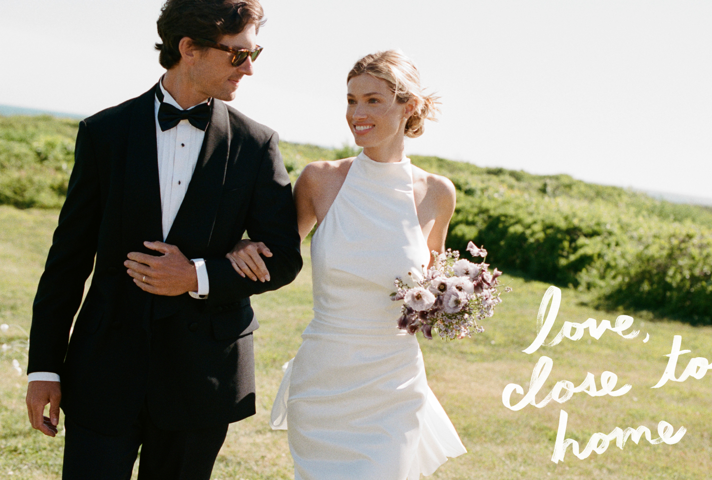 Bride in white halter dress and groom in black tuxedo walking arm-in-arm outdoors with pink bouquet on sunny day