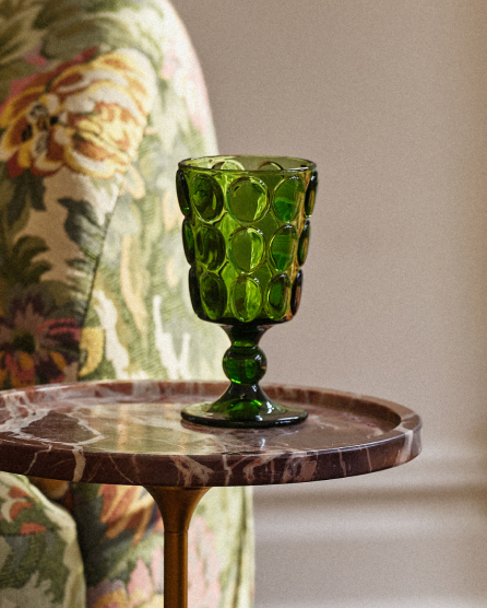 Green textured glass goblet on a round wooden side table next to a floral patterned armchair against a beige wall.