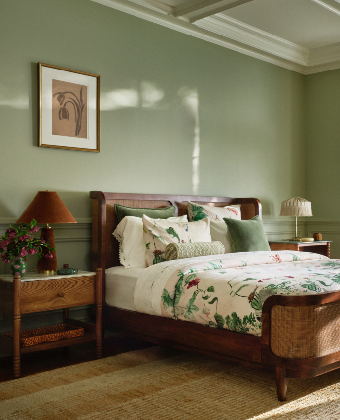 Bedroom with sage green walls, wooden bed with botanical print bedding, green pillows, framed art, and bedside lamps.