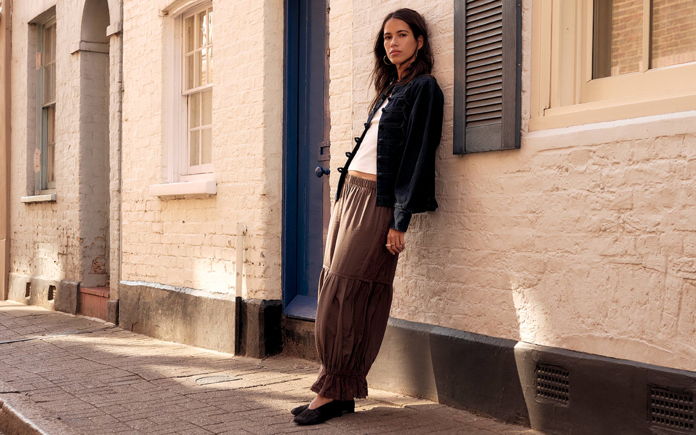 Woman in black denim jacket and brown tiered maxi skirt leaning against cream brick building with blue door and shutters.