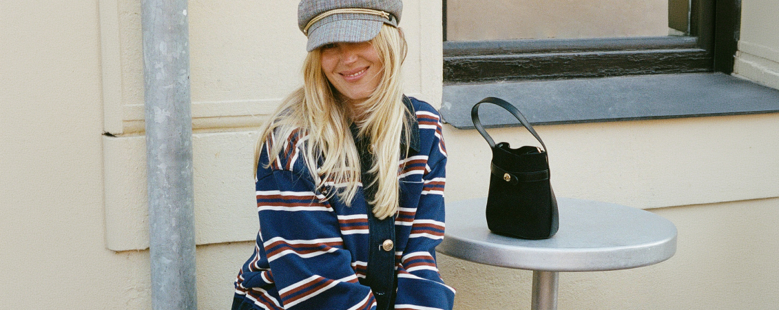Smiling blonde woman wearing a gray cap and navy striped sweater sits at an outdoor table with a black handbag beside her.