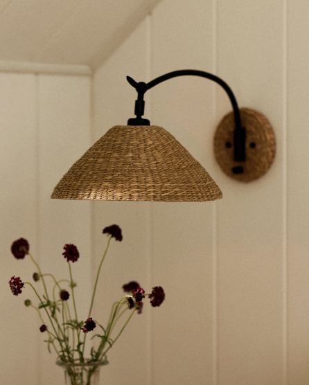 Woven rattan pendant lamp with black metal arm mounted on white paneled wall above a vase of dark purple flowers.