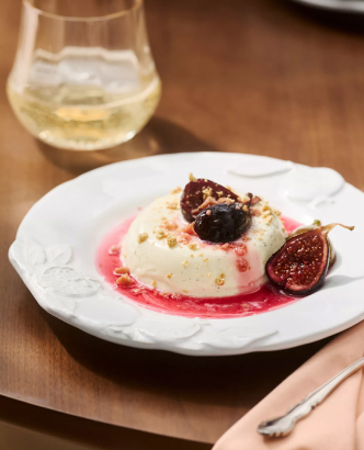 White panna cotta dessert topped with roasted figs and red berry sauce on a white plate, with a glass of white wine nearby.