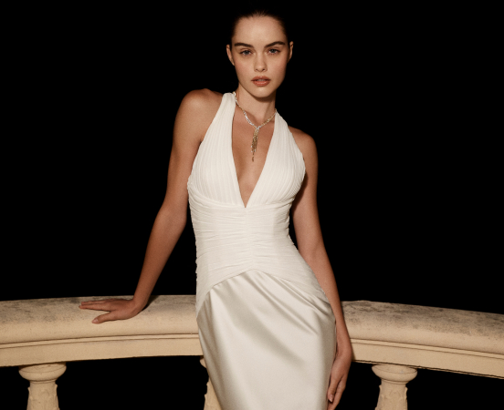 Woman in elegant white halter dress with deep V-neck and pendant necklace, seated on stone balustrade against black background