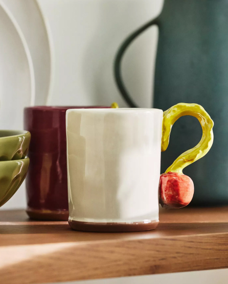 White ceramic mug with unique yellow question mark-shaped handle tipped in red, displayed with other colorful mugs on wooden surface.