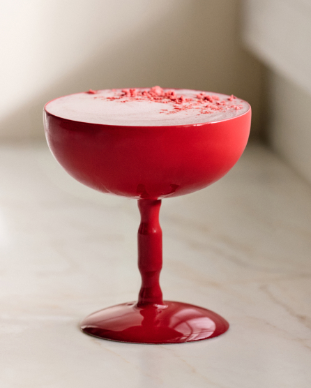 Red coupe glass filled with a pink cocktail topped with white foam and red sprinkles on a marble surface.