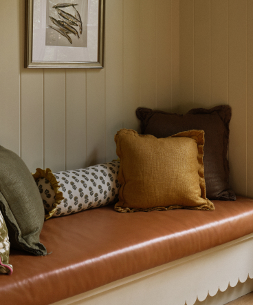 Brown leather bench seat with mustard yellow, brown, and patterned cushions against cream paneled wall with framed artwork above.
