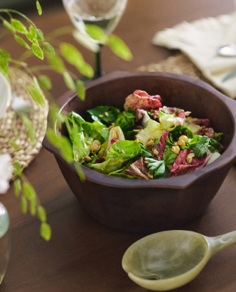 Fresh salad with mixed greens and toppings in a dark wooden bowl on a dining table with greenery and place settings