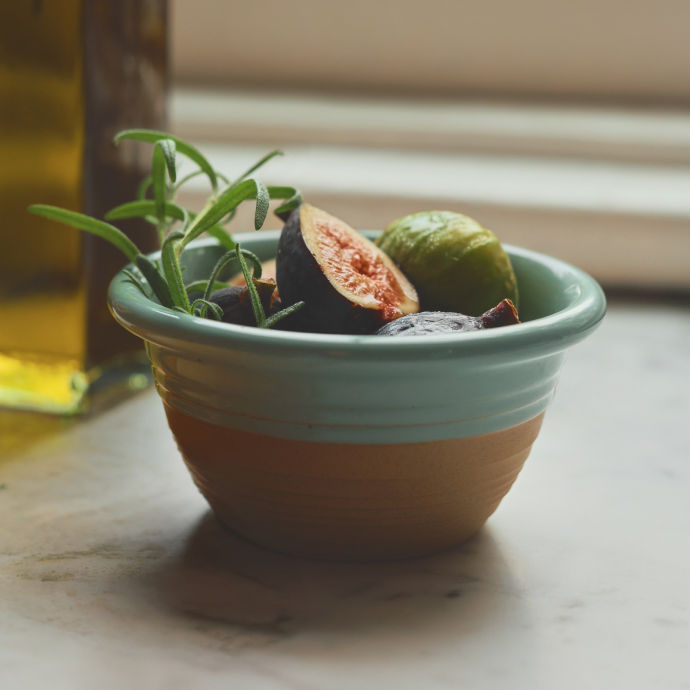 Turquoise and tan ceramic bowl containing fresh figs, one sliced open showing pink flesh, with green herbs and olive oil bottle nearby.