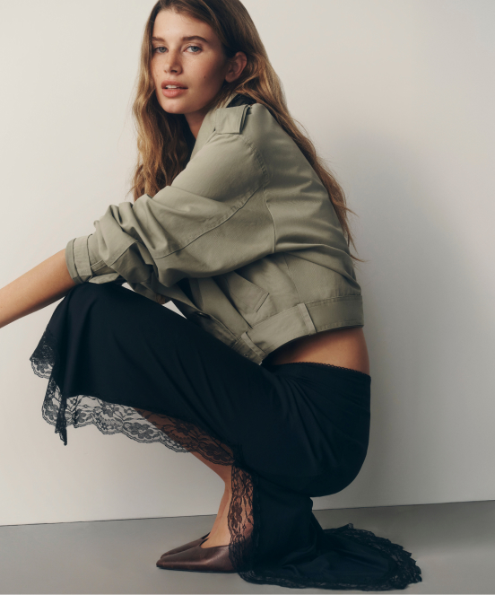 Woman crouching in sage green cropped jacket, black lace-trimmed pants, and brown flats against white background.