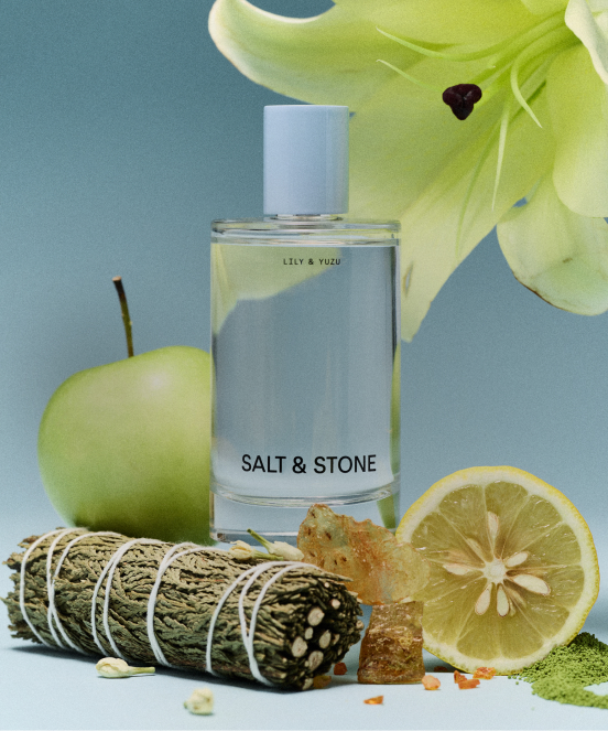 SALT & STONE perfume bottle labeled LILY & YUZU with green apple, lemon slice, sage bundle, and lily flower on blue background.