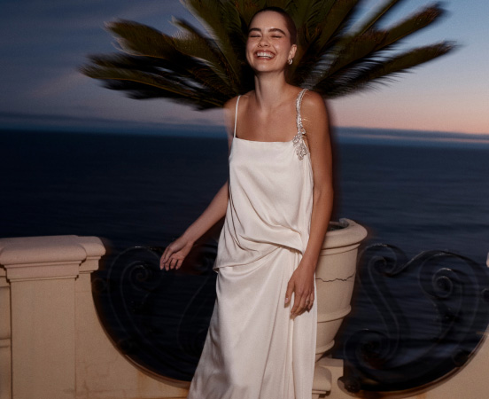 Woman in white dress laughing with hair flowing on ocean balcony at dusk. Text reads BOOK AN APPOINTMENT.