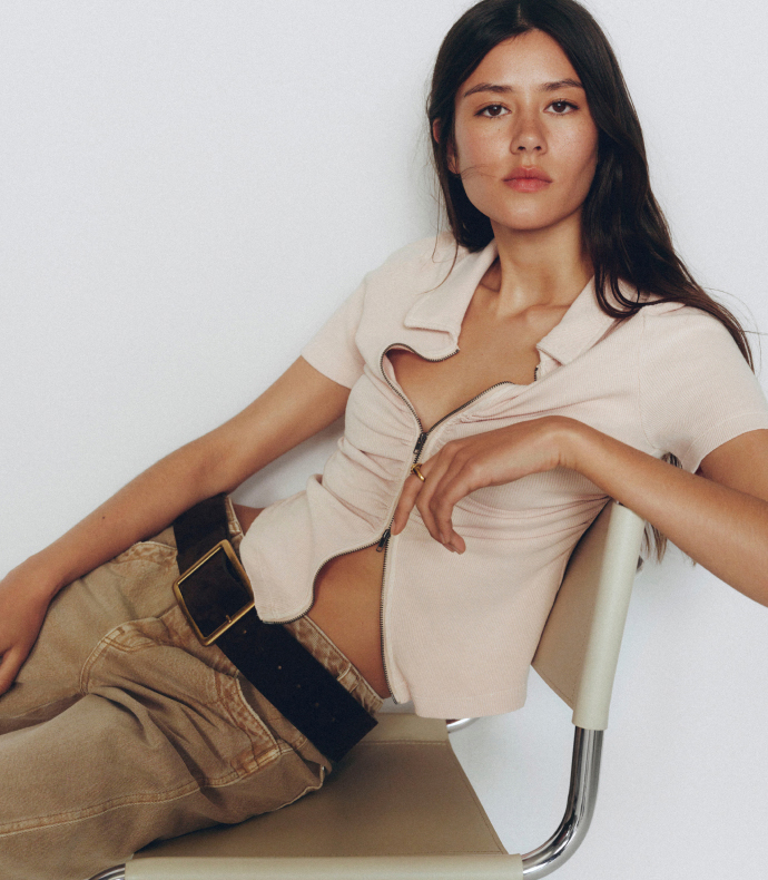 Woman with long dark hair sitting on white chair wearing beige polo shirt and tan pants with black belt against white wall.