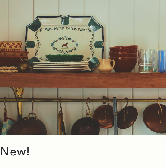 Decorative tray with dog motif on wooden shelf above hanging copper pots in rustic kitchen with cream paneled walls.