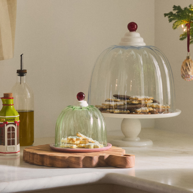 Two glass cake domes with red cherry-topped handles covering cookies on white pedestals, with an olive oil bottle nearby.
