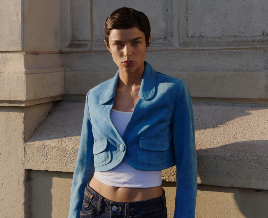Young woman in cropped blue jacket and white top standing against concrete wall with columns in natural sunlight.
