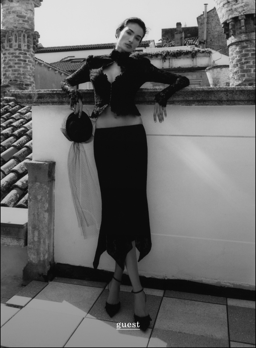 Black and white photo of woman in elegant black outfit with handbag on rooftop terrace with stone towers. Text reads guest.