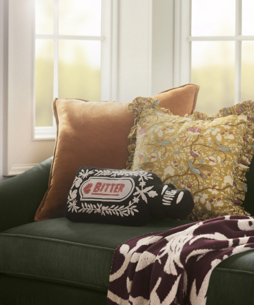 Green velvet sofa with decorative pillows in rust, yellow textured, and black with white embroidered design near sunny window.