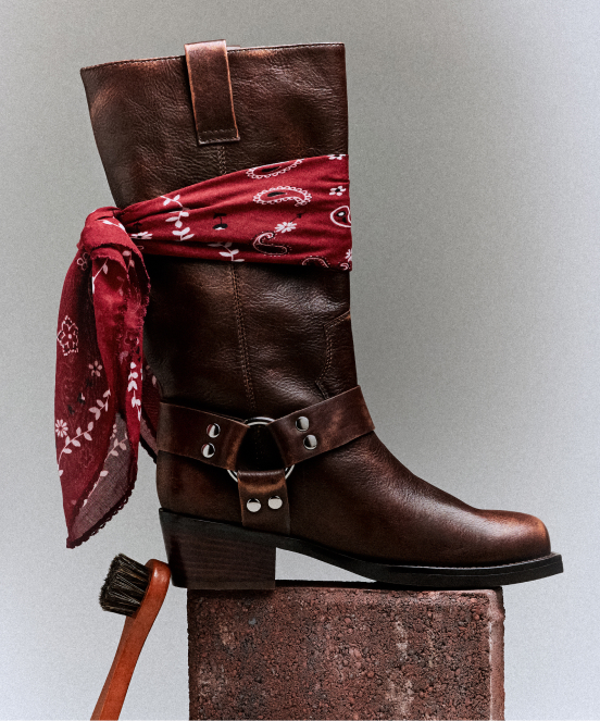 Brown leather boot with red paisley bandana tied around shaft and metal harness detail, displayed on rustic block with brush.