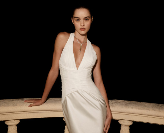 Model in elegant white halter wedding gown with deep V-neck and ruched bodice, seated on beige bench. Text reads "WEDDING GOWNS"