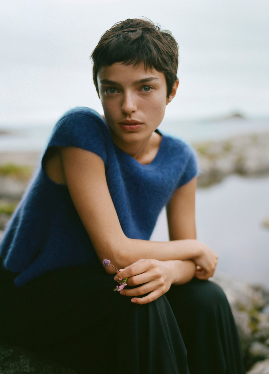 Woman with short dark hair in blue shirt sitting outdoors by water, holding small purple flowers, looking at camera.