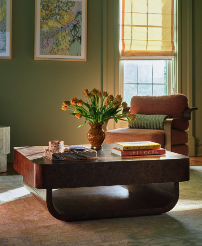 Modern living room with curved dark wood coffee table, orange tulips in vase, stacked books, and rust-colored armchair by window.