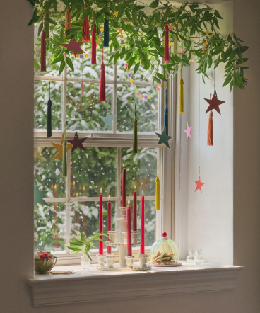 Window decorated with hanging red candles, greenery, and star ornaments with a white windowsill display below.