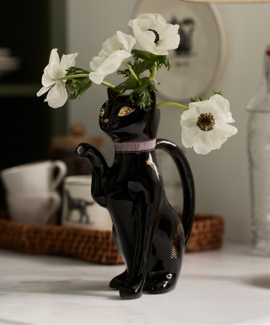 White anemone flowers in a black cat-shaped ceramic vase with gold eyes and pink collar on a white table.
