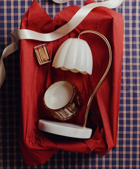 Vintage brass lamp with white glass shade, matchbox, and white plate arranged on red fabric over blue plaid background.