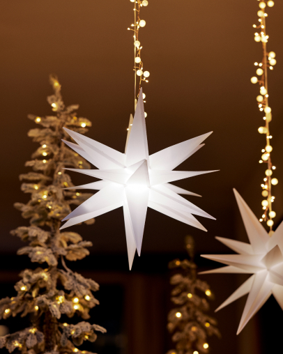 White illuminated star ornament hanging with golden Christmas tree and string lights in background against brown wall.
