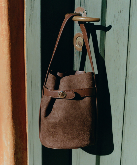 Brown suede bucket bag with leather strap hanging on a weathered green door handle with keyhole visible.