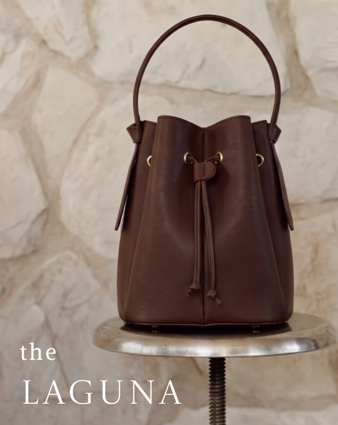 Brown leather drawstring bucket bag labeled "the LAGUNA" displayed on a marble pedestal against a white fabric backdrop.