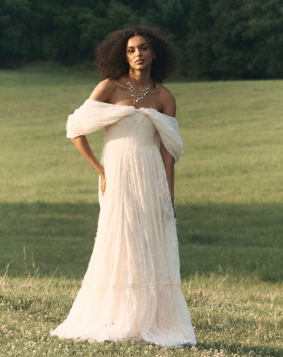 Woman in white off-shoulder gown with statement necklace standing in a sunlit field with green trees in background.