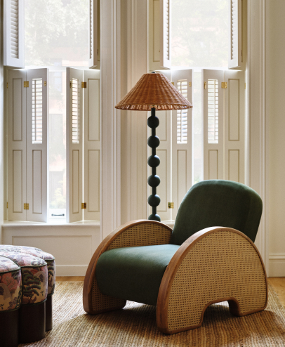 Green velvet chair with woven rattan arms beside a tall floor lamp with black spherical base and wicker shade in a bright room with shuttered windows.