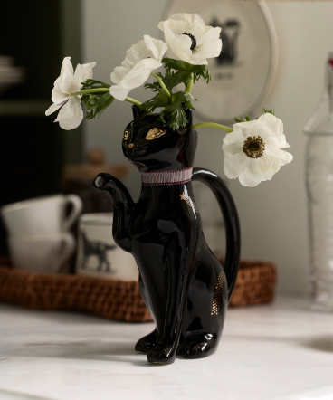 White anemone flowers arranged in a black cat-shaped vase with gold accents on a white surface with wicker basket behind.
