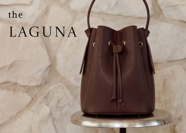 Brown leather drawstring bucket bag labeled "the LAGUNA" displayed on a marble pedestal against a white fabric backdrop.
