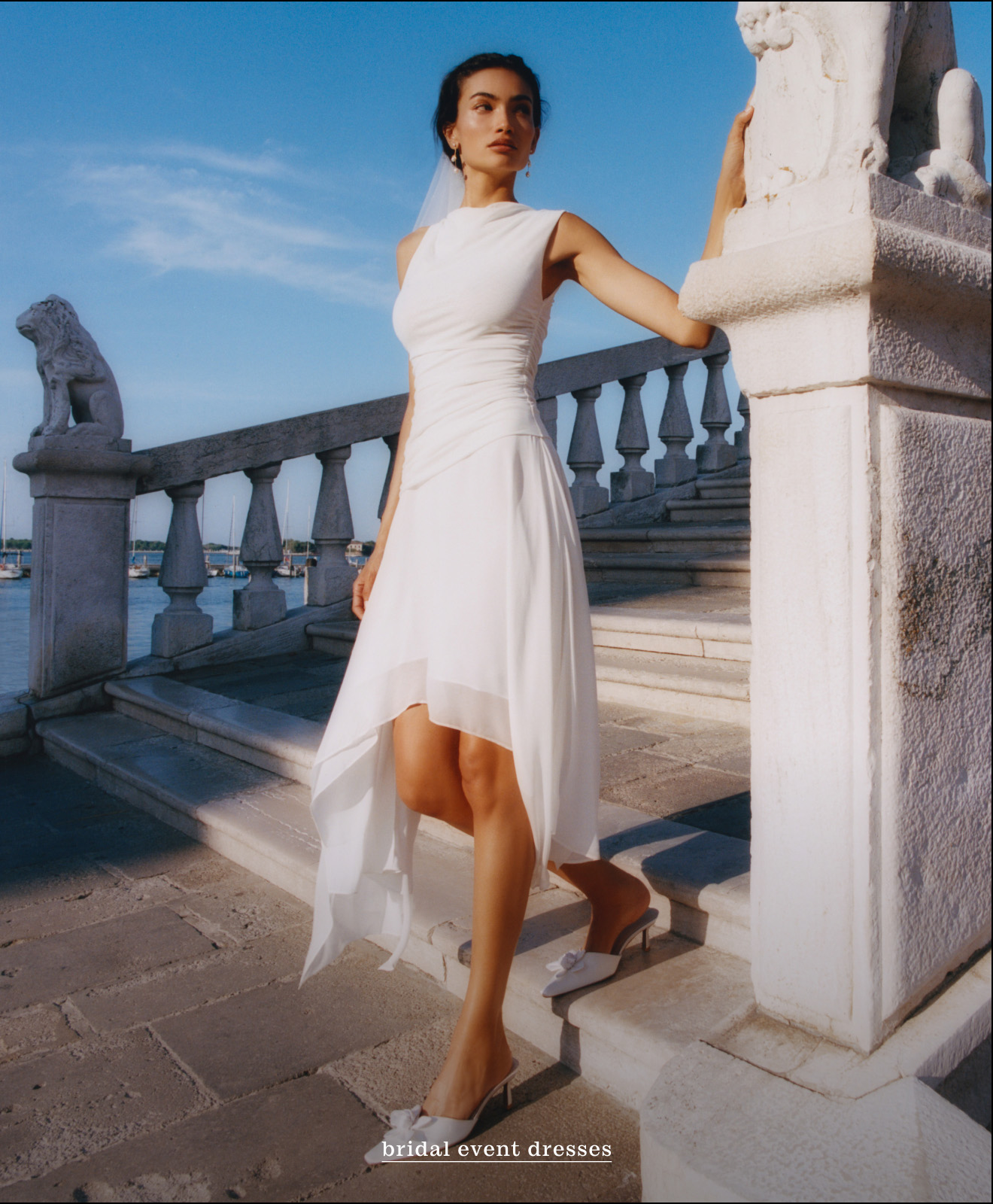 Woman in white asymmetrical dress on stone terrace with balustrade and lion statue against blue sky and water backdrop.