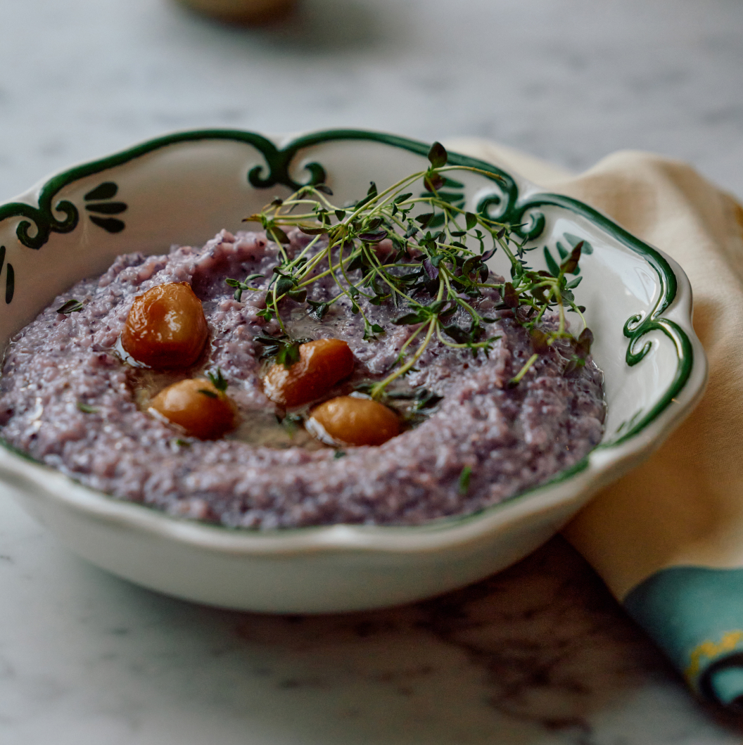 Purple hummus topped with chickpeas and fresh thyme in a decorative white bowl with green trim on a marble surface.