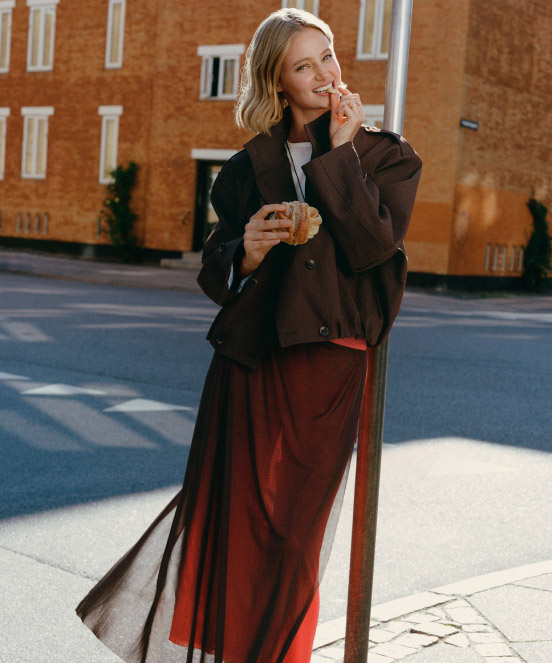 Woman in brown jacket and rust-colored skirt eating pastry and drinking coffee outdoors with brick building behind her.