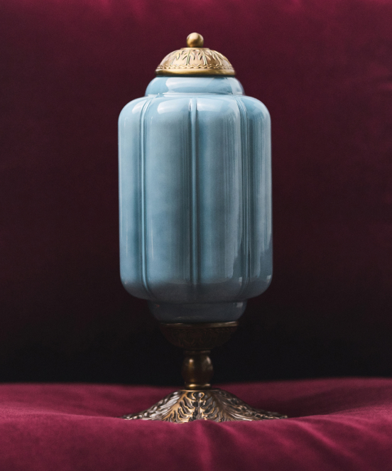 Light blue ceramic jar with ornate brass lid and decorative brass base on burgundy velvet background