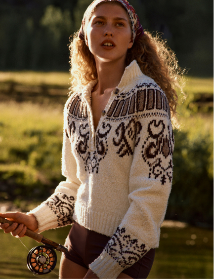 Young woman with curly hair wearing a patterned white and brown zip-up sweater outdoors in natural sunlight.