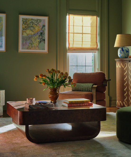 Green-walled living room with modern dark wood coffee table, orange tulips in vase, brown armchair, and yellow roman shade on window.
