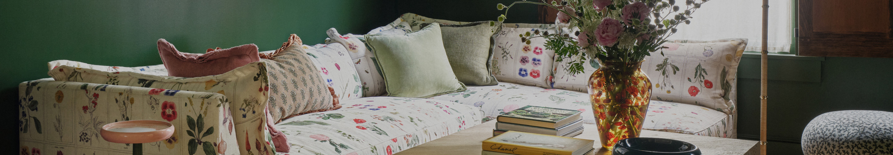 Floral-patterned sofa with mixed cushions, decorative vase with flowers, stacked books on glass table against green wall.