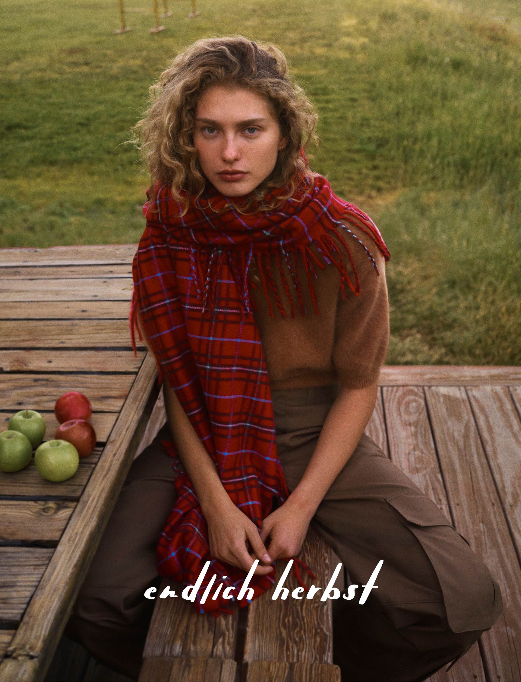 Woman in brown sweater and olive pants on wooden deck with mountain landscape, text reads "autumn, at last"