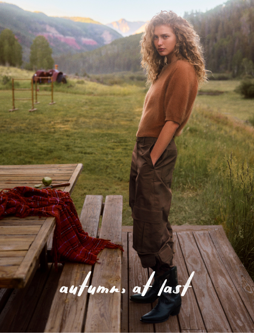 Woman in brown sweater and olive pants on wooden deck with mountain landscape, text reads "autumn, at last"