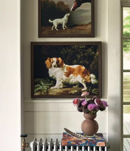 Two framed vintage dog paintings hang on a white wall above a console table with a vase of purple flowers and stacked books.