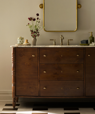 Dark wood bathroom vanity with marble top, gold-framed mirror, and floral arrangement against beige wall