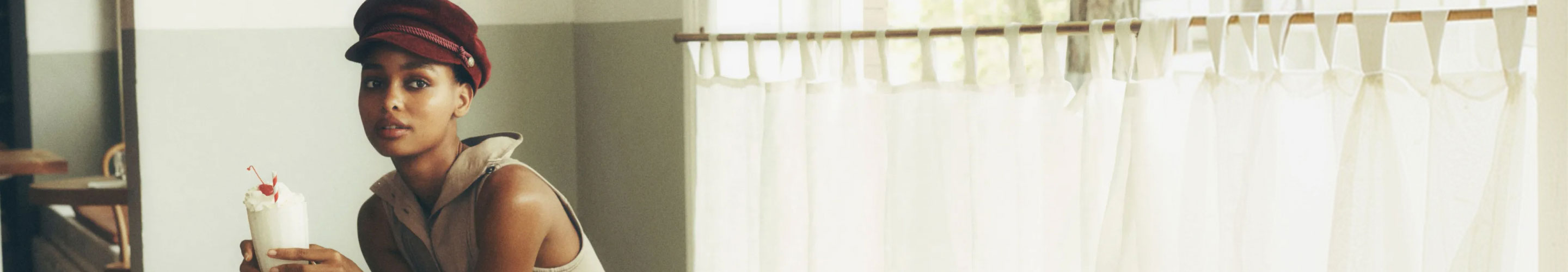 Woman wearing burgundy cap holds white beverage cup with straw in bright interior space with white curtains