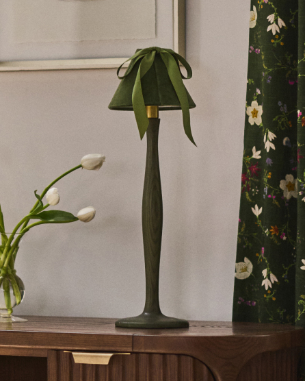 Green palm leaf-shaped table lamp on wooden surface with white tulips in vase and floral curtain in background.