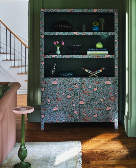 Floral-patterned bookshelf with black shelves displaying decorative items, set in a green-framed alcove near a staircase.
